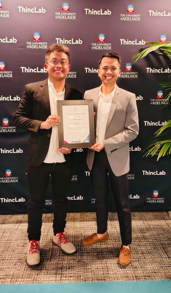 Gerald and PJ smile proudly holding a framed certificate for first prize. They stand in front of a navy University of Adelaide ThincLab banner wearing white shirts, dark pants and suit jackets, with a palm on the side. 
