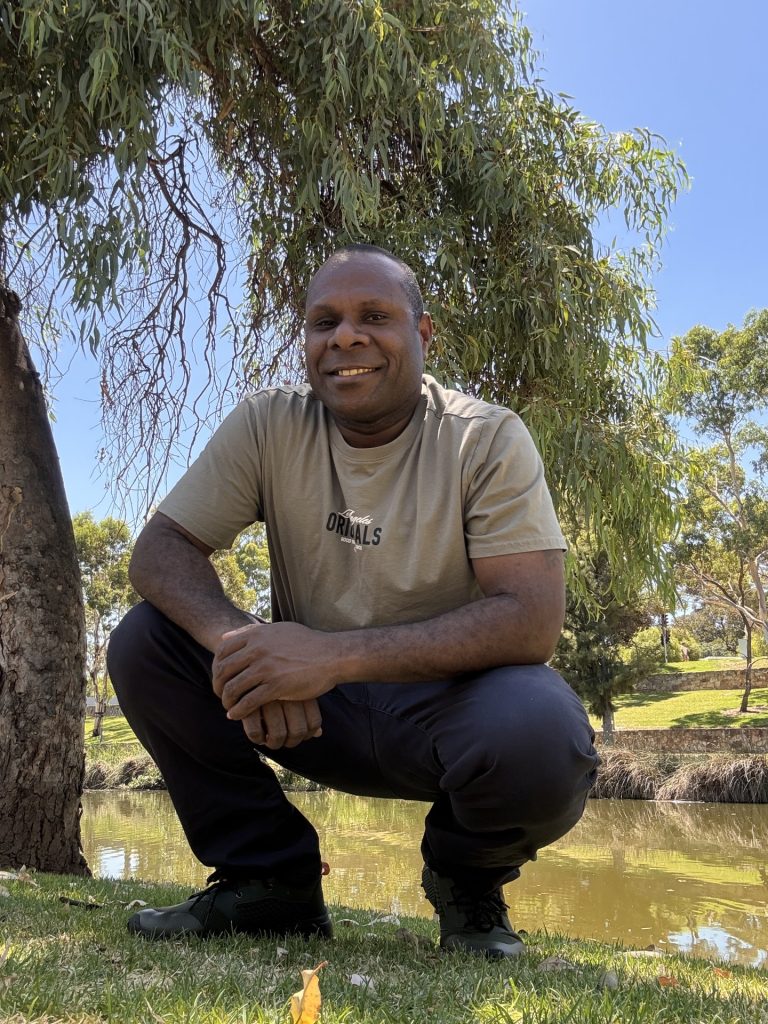 Sailas crouches on the grass next to a tree in front of a river. He smiles and wears a t-shirt and jeans.