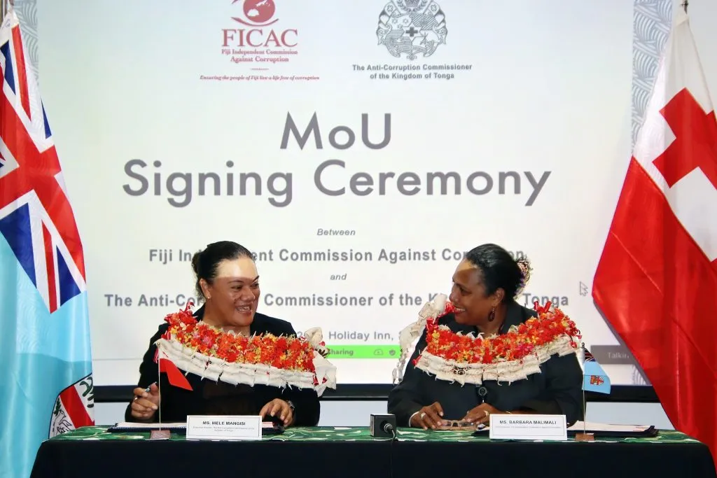 Mele and another woman sit at a desk together smiling at each other while signing papers. A sign behind them says: MoU Signing Ceremony with Tonga and Fiji flags beside them