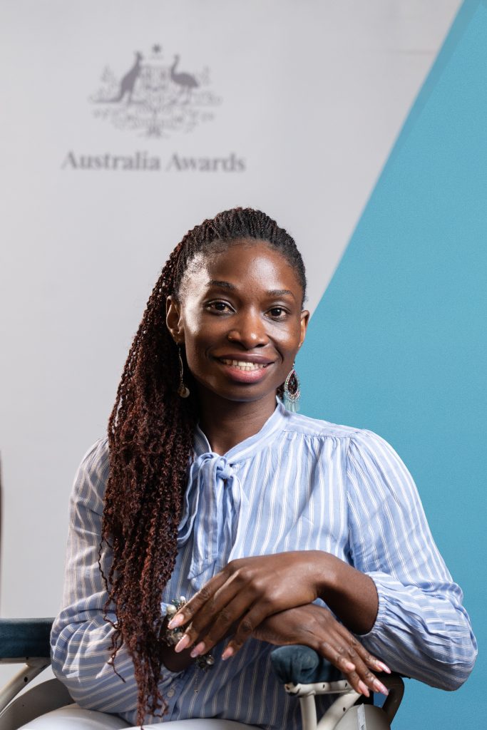 Victo is smiling and sits in a wheelchair wearing a light blue striped blouse. Behind her is the Australia Awards backdrop.