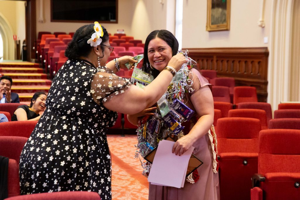 Esterlina is presented with a lei containing money, on top of leis with chocolate and ribbons, by a family member. She holds her graduation certificate and is smiling.