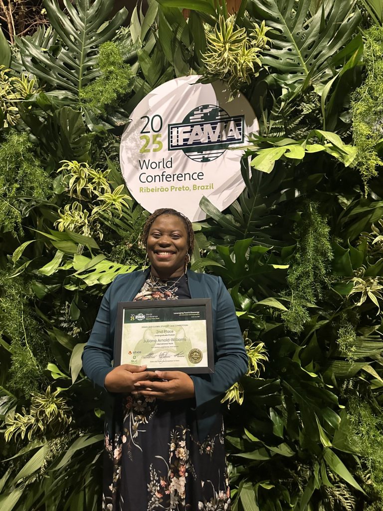 Julianna is smiling and holding a certificate. She stands in front of a very lush green background of leaves with a 2025 IFAMA World Conference Brazil sign. 