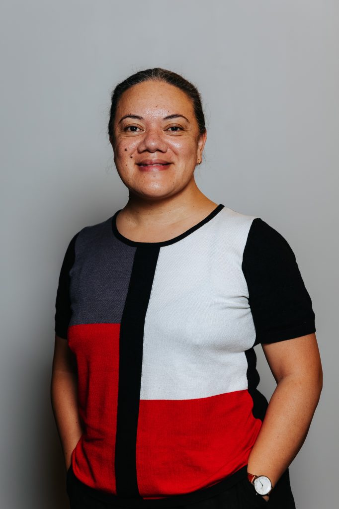 Mele smiles with her hands in her pockets and wears a black, white, red and blue top.