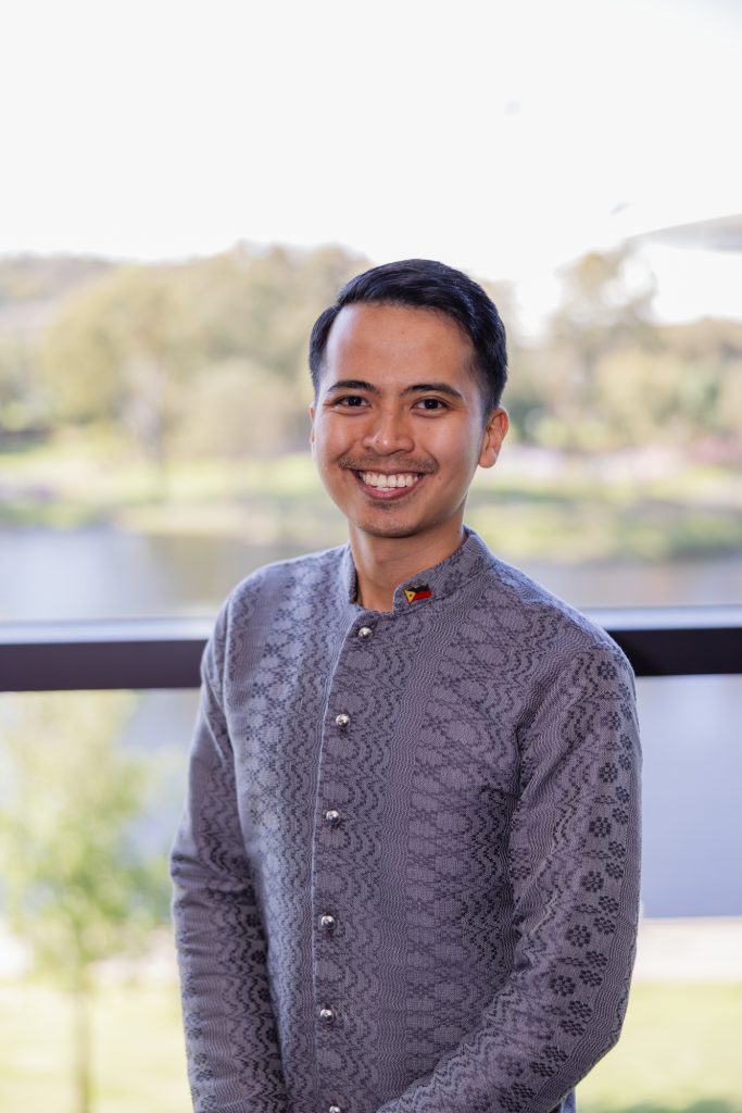 Paul stands smiling in front of a window with the River Torrens behind him. He wears a traditional grey buttoned jacket with a Philippines flag pin.