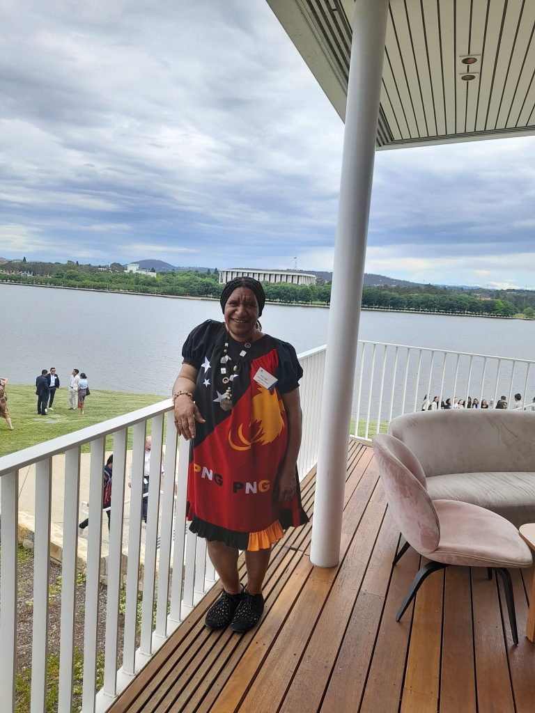 Ruth is smiling and standing on a wooden deck with white railings, wearing a long meri blouse in traditional PNG colours and patterns. There is a group of people standing on the lawn next to Lake Burley Griffin in the background and the National Library can be seen across the lake.
