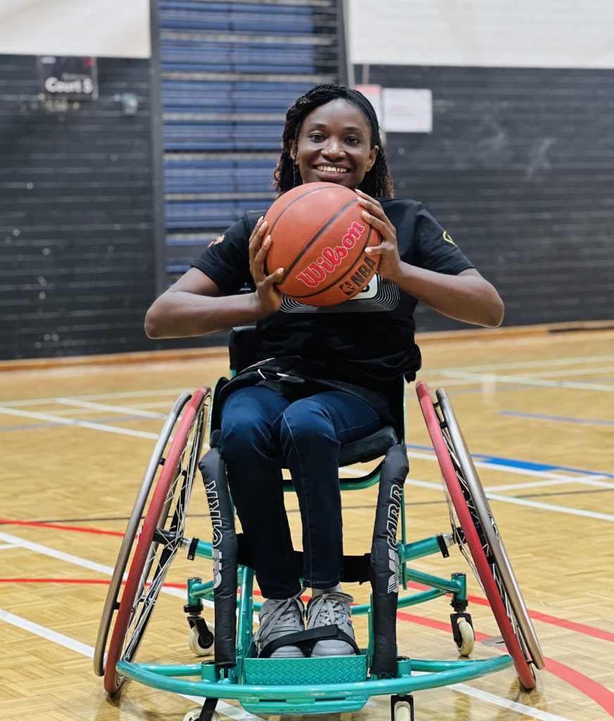 Victo sits in a wheelchair adapted for basketball smiling broadly and holding a basketball. 
