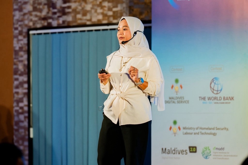 Moomina is speaking on stage wearing a headset microphone, a white hijab and jacket with text: World Public Service Day.