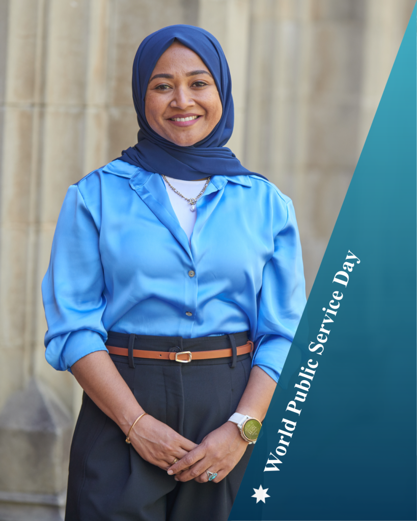 Moomina stands in front of sandstone columns wearing a blue shirt, navy pants and blue hijab smiling. Text: World Public Service Day.