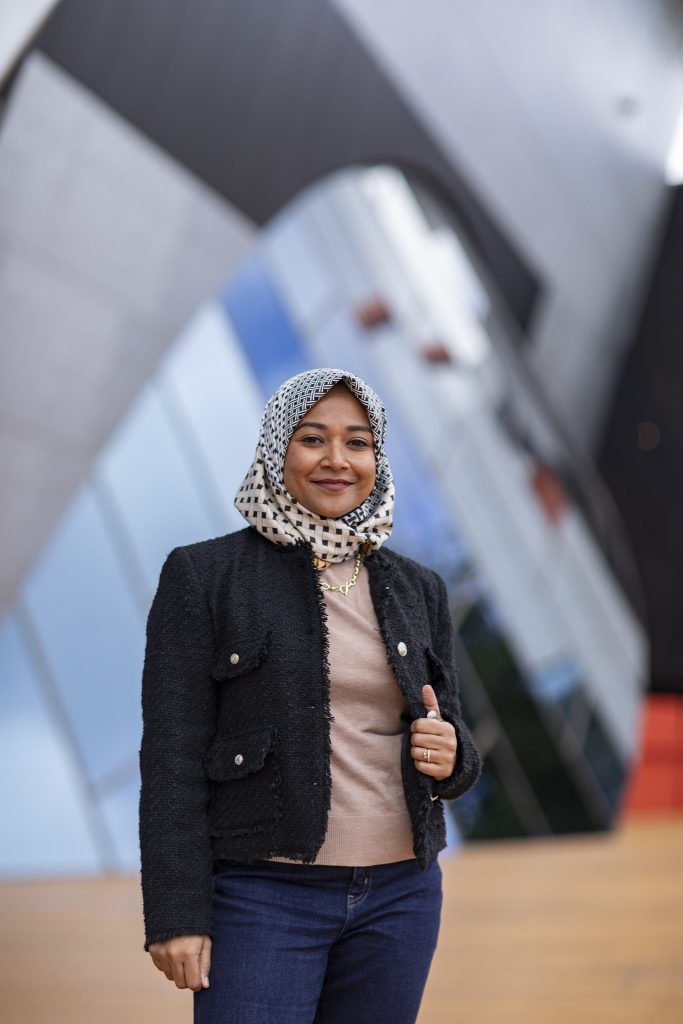Moomina stands outside smiling in casual clothing in front of a building with sloping walls and a reflective glass feature.