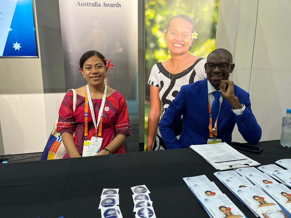 A female and male scholar sit in the Australia Awards booth with banners and a screen behind them and Australia Awards stickers and pamphlets on the table in front of them.