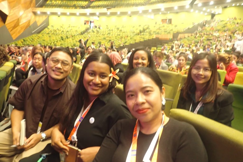 Participants sit smiling in a large auditorium wearing lanyards. 
