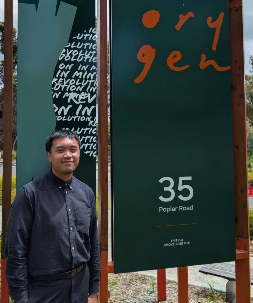 Ash stands in front of the Orygen sign outside smiling and wearing a black shirt and pants.