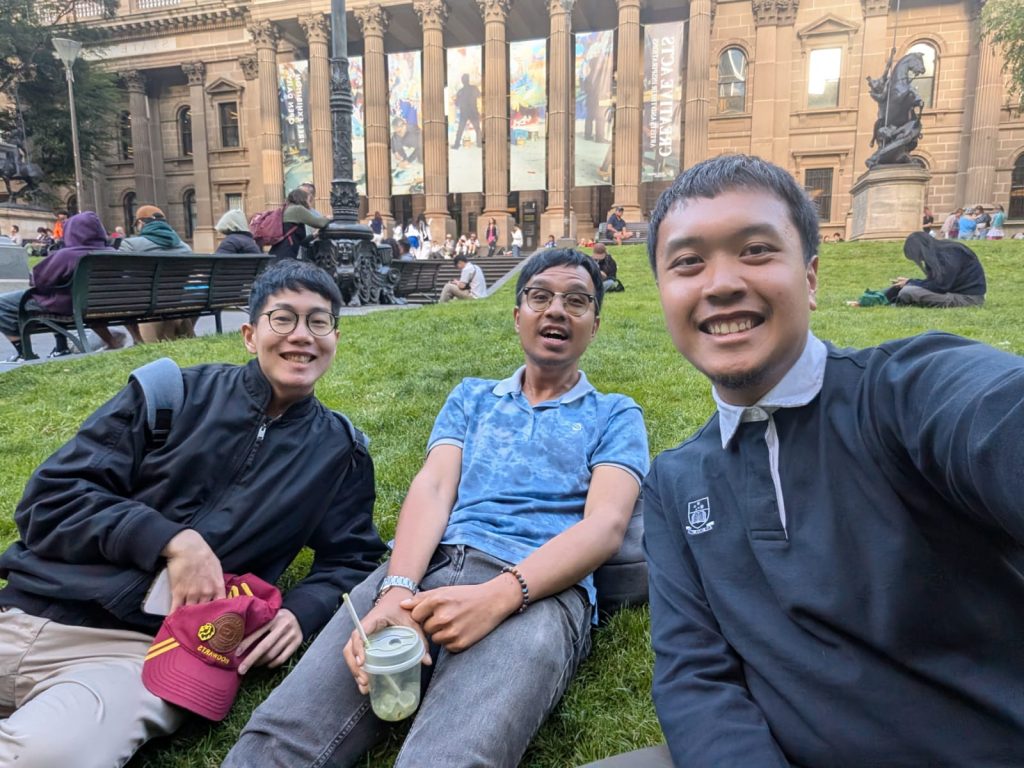 Ash sits on the lawn with two other men in front of the Melbourne State Library.