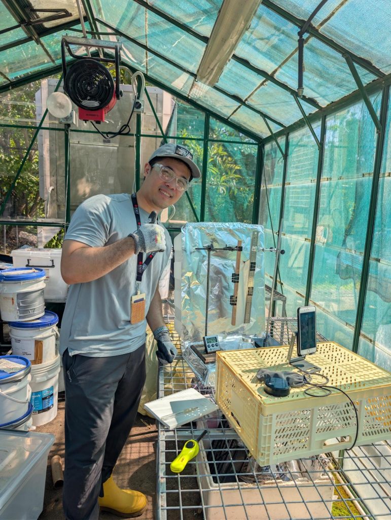 Arekha stands in a lab smiling wearing jeans, t-shirt, gloves and cap using machines connected to a mobile phone with large plastic tubs behind him.