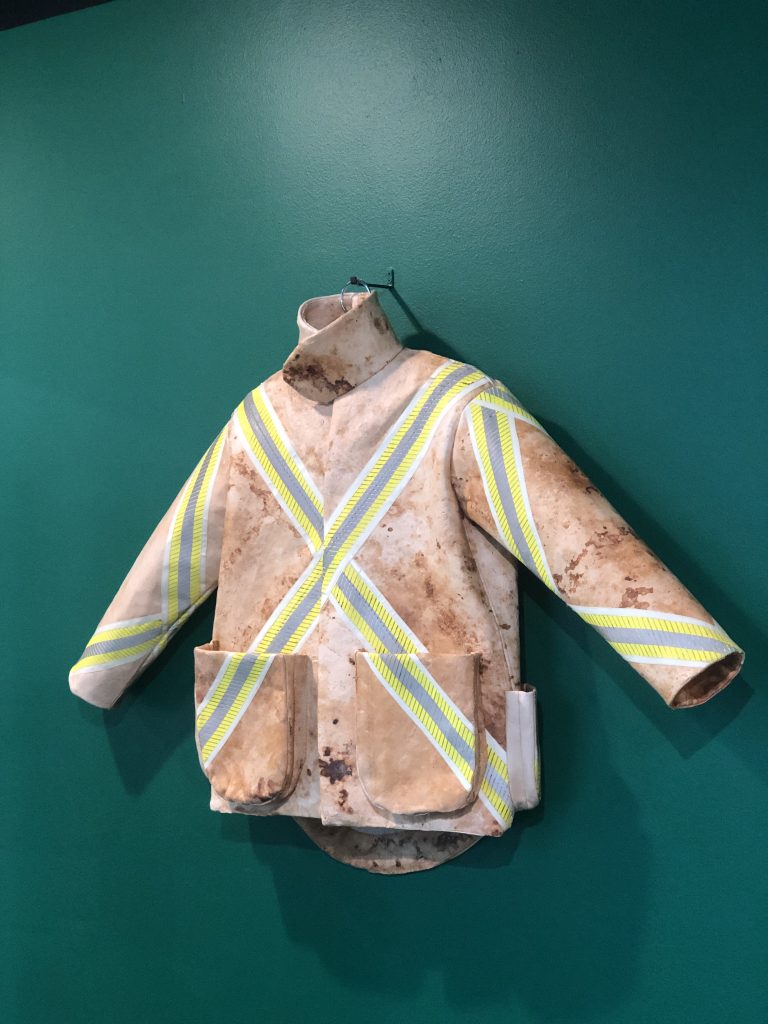 A light coloured jacket that appears to have natural marks like dirt, and a reflective cross on the front is pinned to a green wall on display.