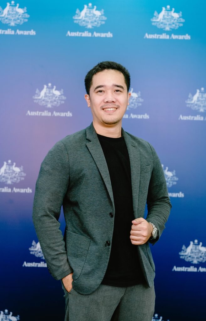 Arekha stands smiling in a grey suit jacket and trousers with a black top in front of an Australia Awards media wall.