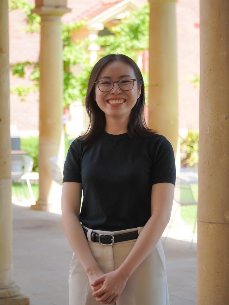 Li Ling stands between columns at Adelaide University smiling and wearing glasses, a black t-shirt and beige trousers.