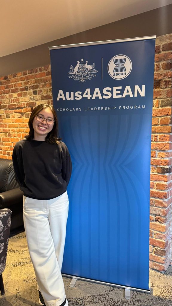 Li Ling stands smiling next to an Aus4ASEAN banner wearing glasses, a black top and white trousers. 
